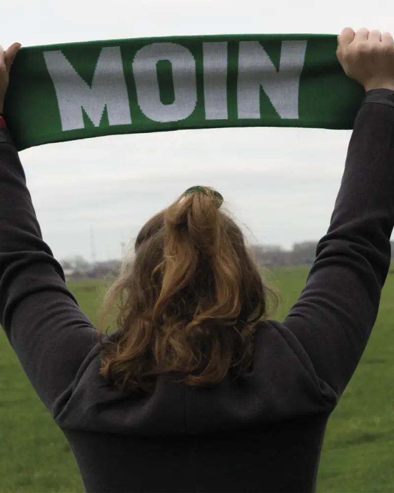 Grüner Fanschal mit der Aufschrift „Moin“, hochgehalten vor einer Wiese